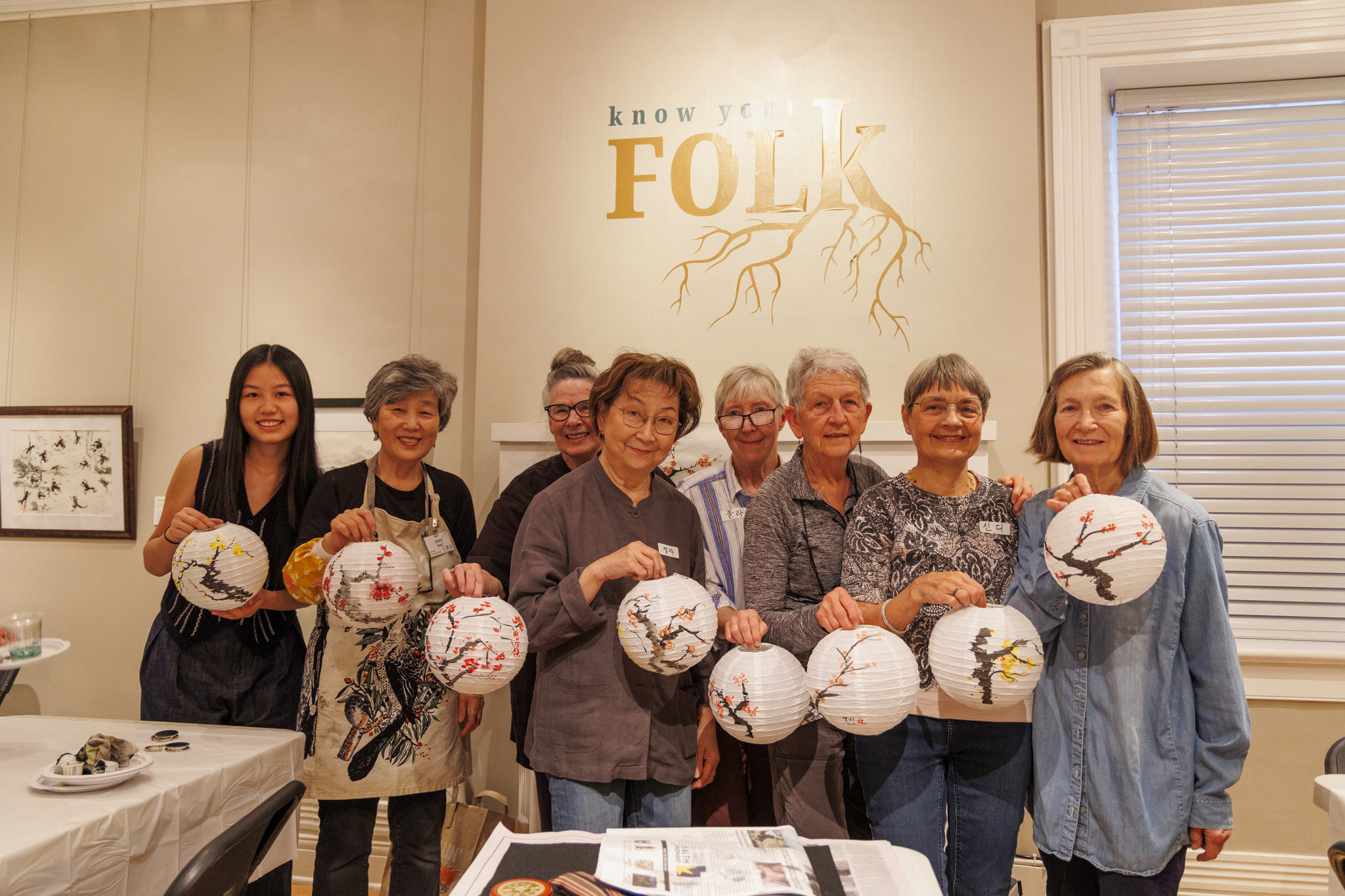A group of eight smiling people holds decorated paper lanterns indoors. Behind them, “Know Your Folk” is written on the wall. The mood is joyful and creative.