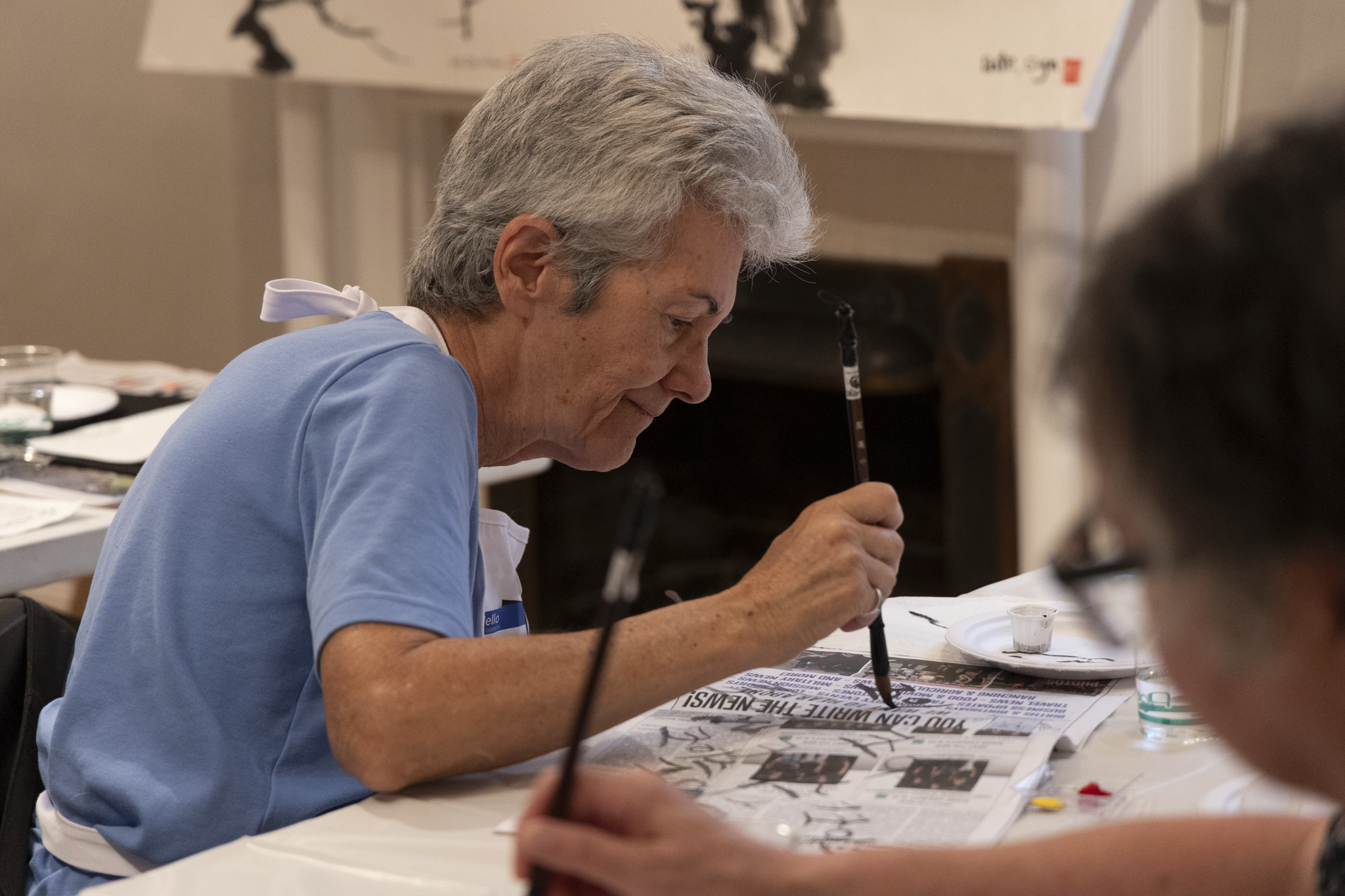 An elderly person with gray hair, in a blue shirt and apron, is focused on painting Chinese characters with a brush on newspaper. The setting is calm and creative.
