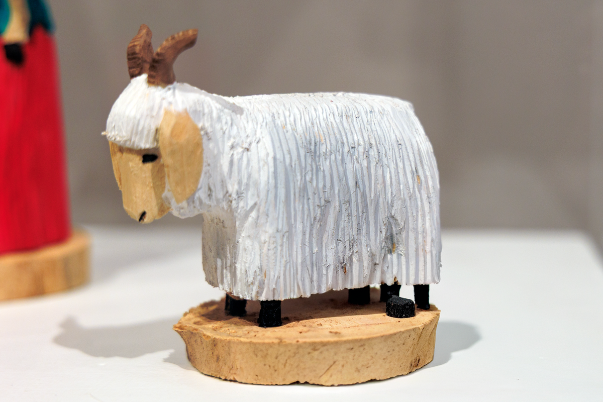 Wooden goat sculpture with white fur texture, brown horns, and a beige face. It stands on a round wooden base. Background is blurred.