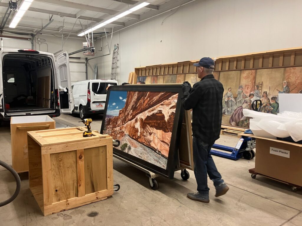 A person moves a large framed painting in a storage area with crates and art supplies.