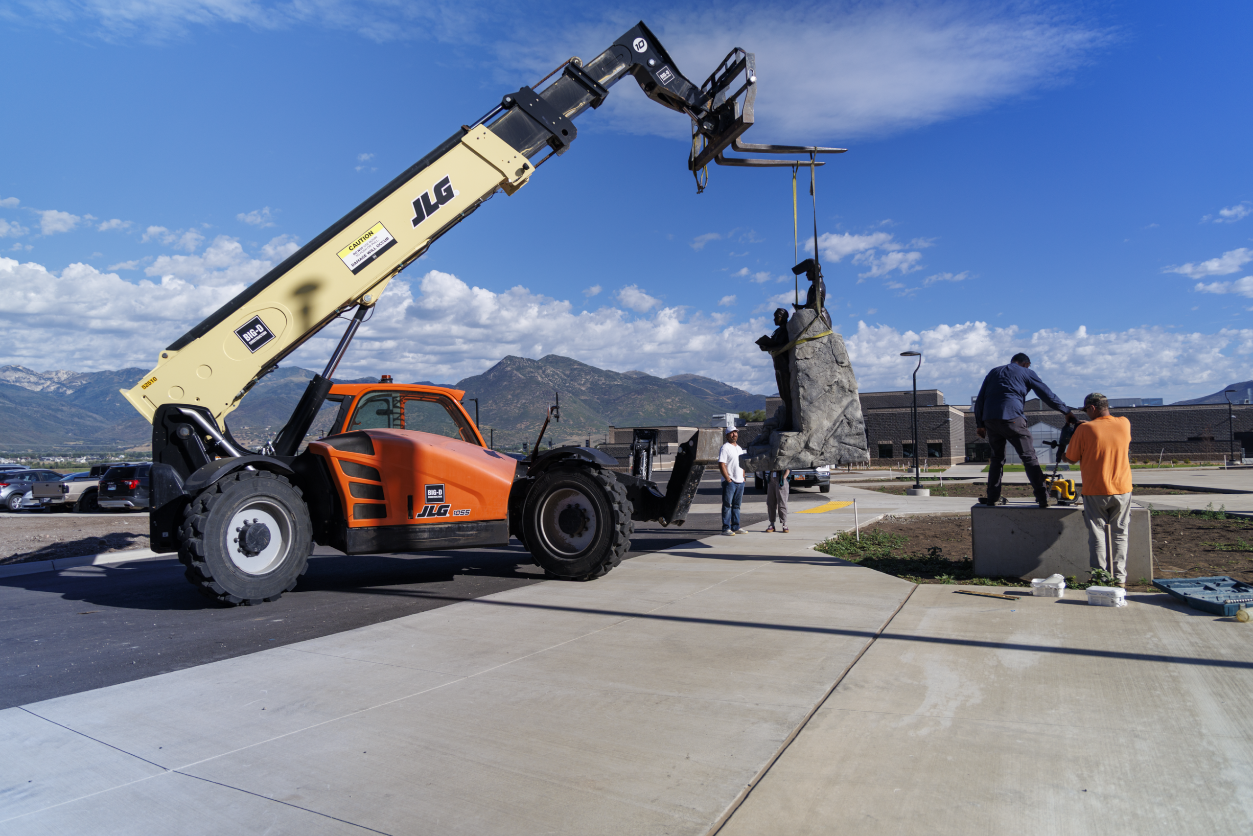 A forklift lifts a statue while several people guide it outdoors on a sunny day.