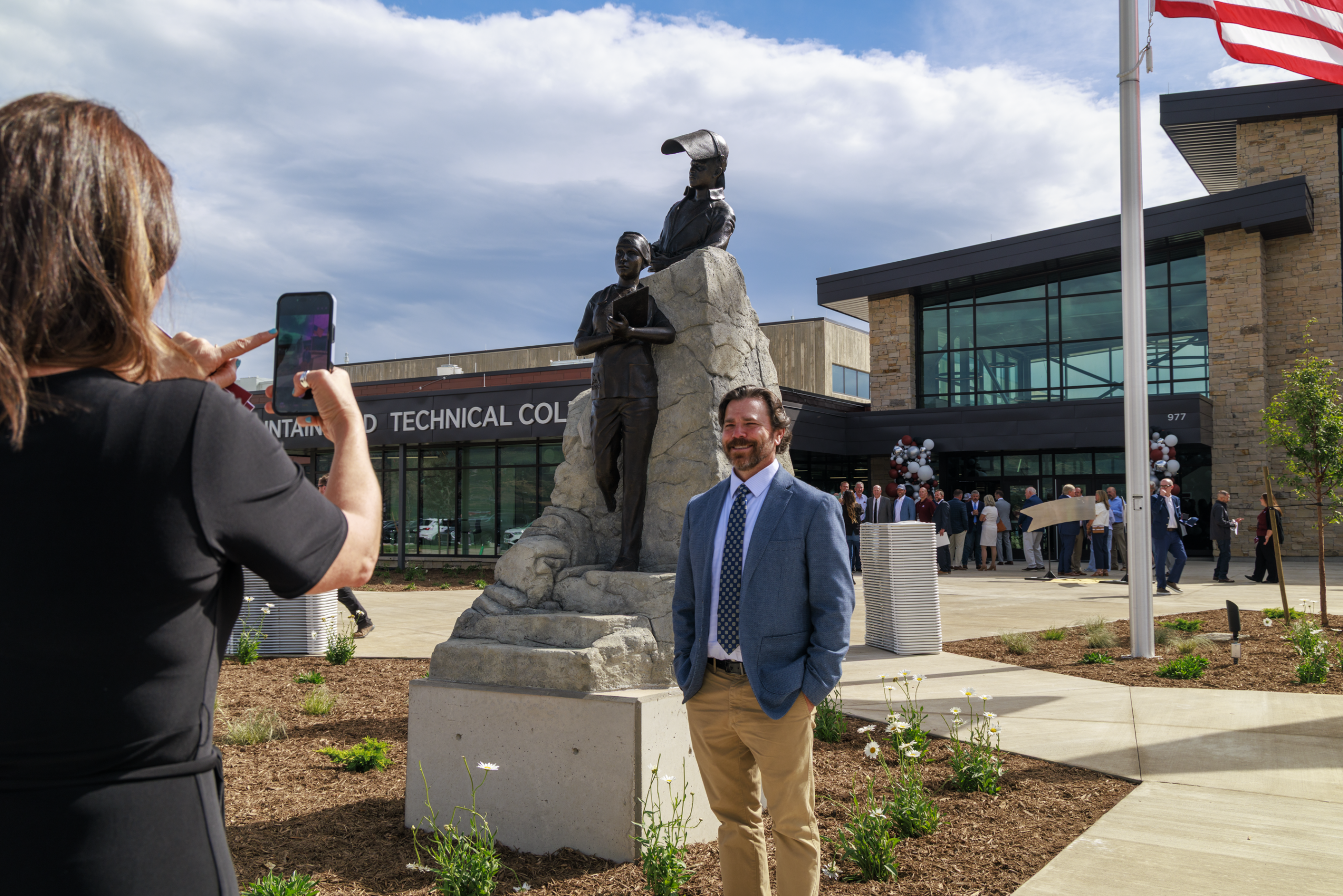 Person poses by statue as another person takes a photo outside a modern building with flags.