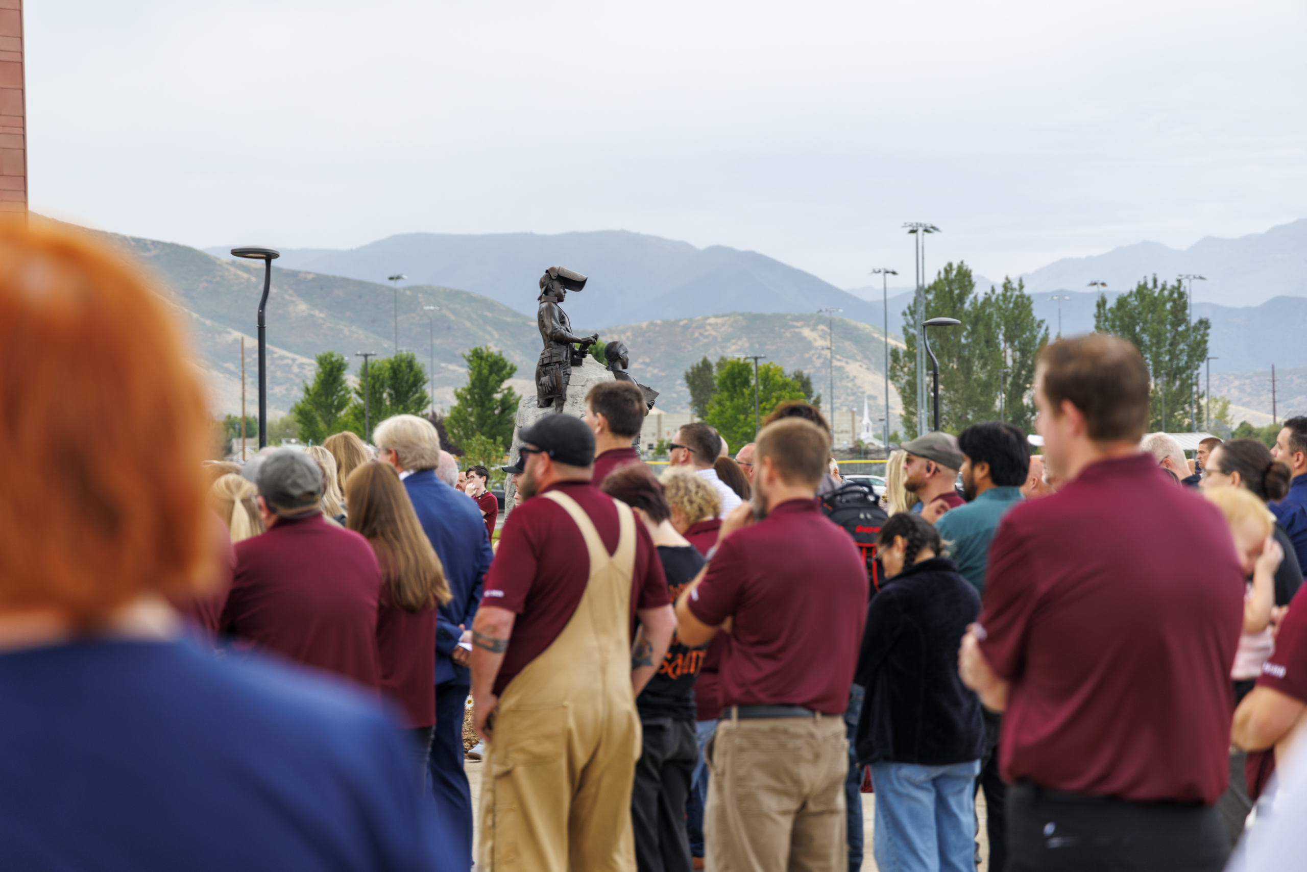 A crowd gathers outdoors near a statue, with mountains visible in the background.