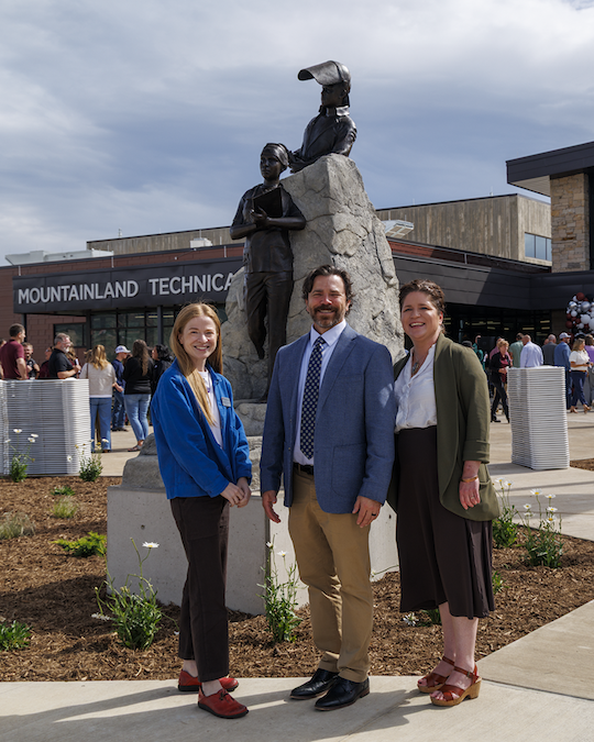 Three people smile in front of a statue at Mountainland Technical College, outdoors.