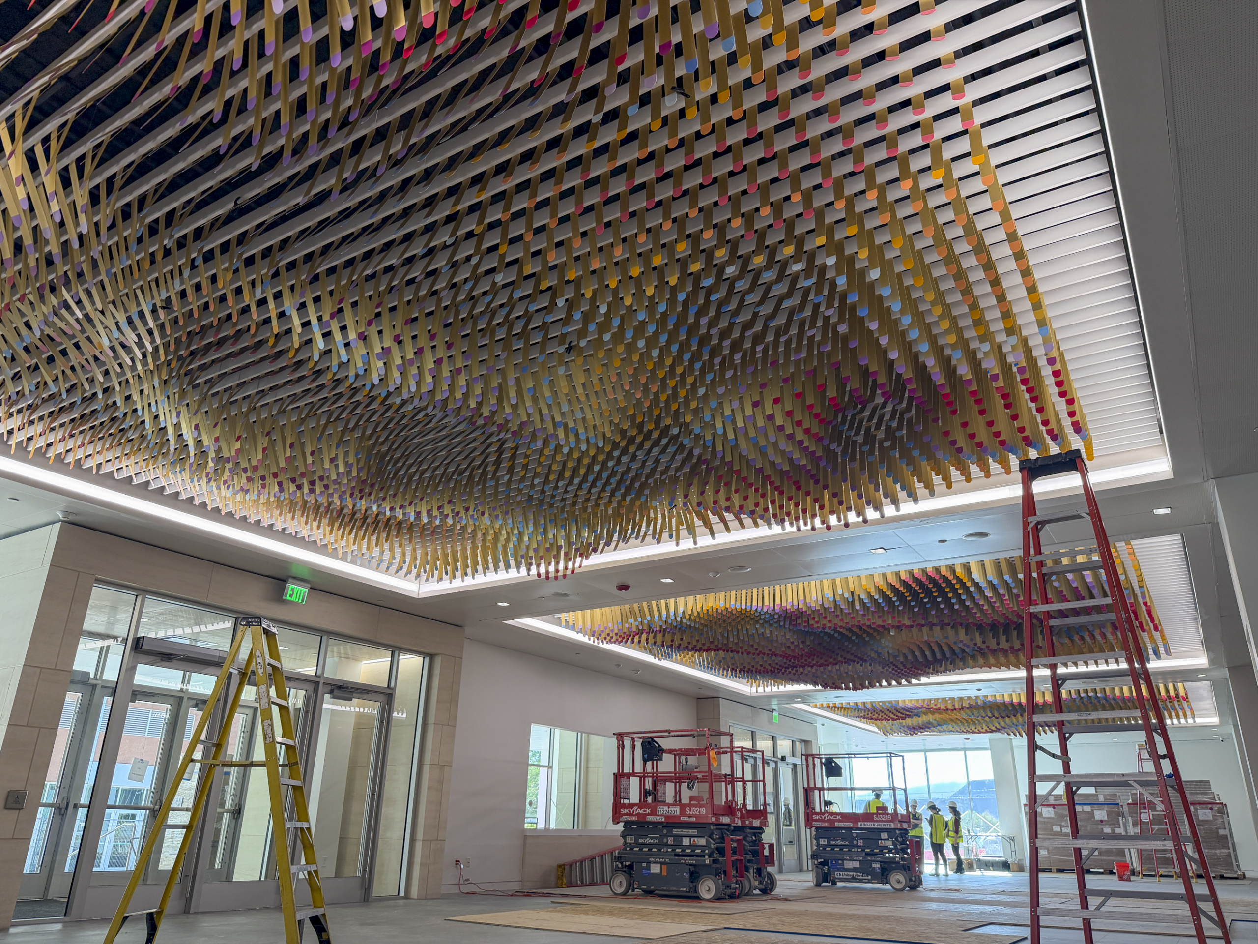 Colorful woven ceiling art installation with ladders and workers in a modern interior space.