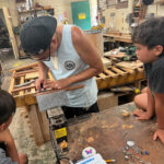 Man working with tools on wood while two children watch in a workshop.